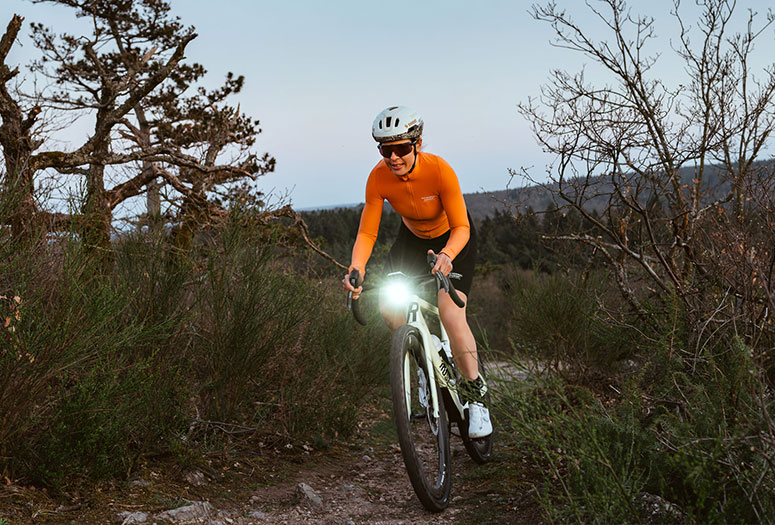 Eine Frau fährt in der Dämmerung abseits befestigter Wege Fahrrad; das helle Licht ihres Rads beleuchtet den Weg vor ihr.
